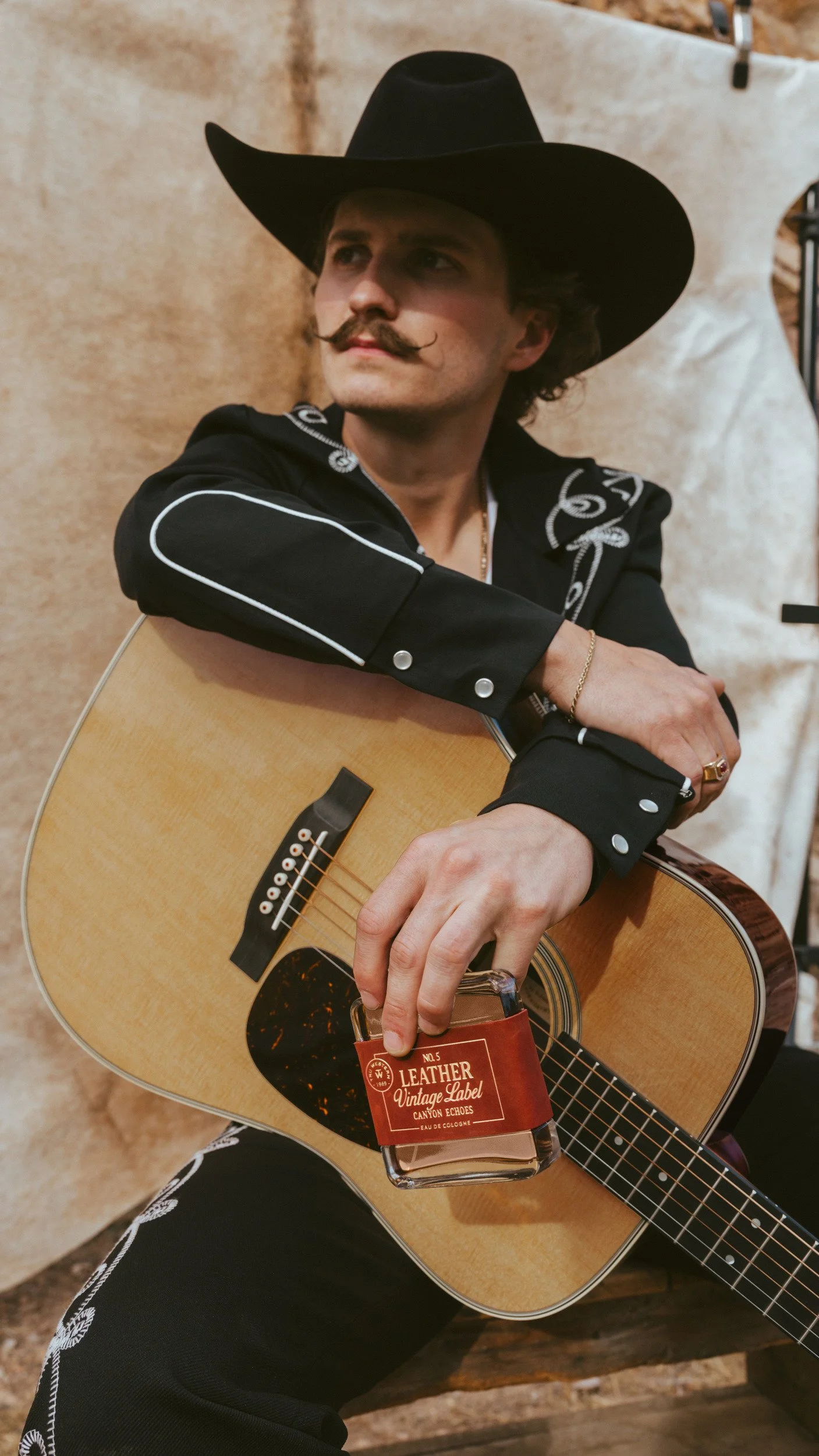 Braxton Keith seated with his guitar at a cowhide backdrop, black hat and embroidered shirt, the bottle held to the guitar.