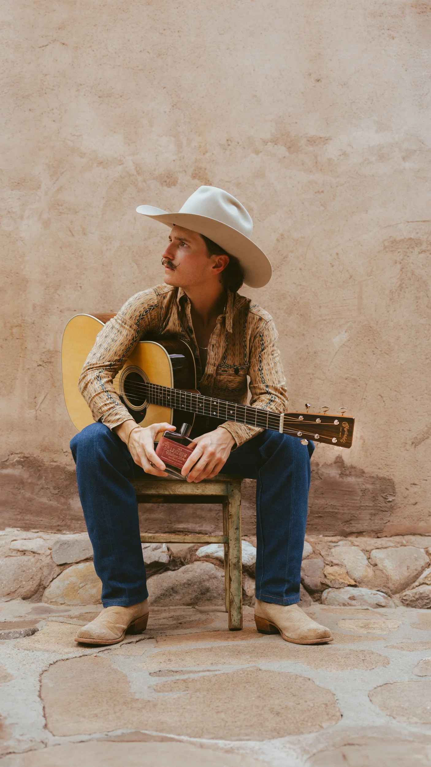 Braxton Keith seated on a wooden chair against an adobe wall, guitar across his lap, white hat, jeans and pale boots.
