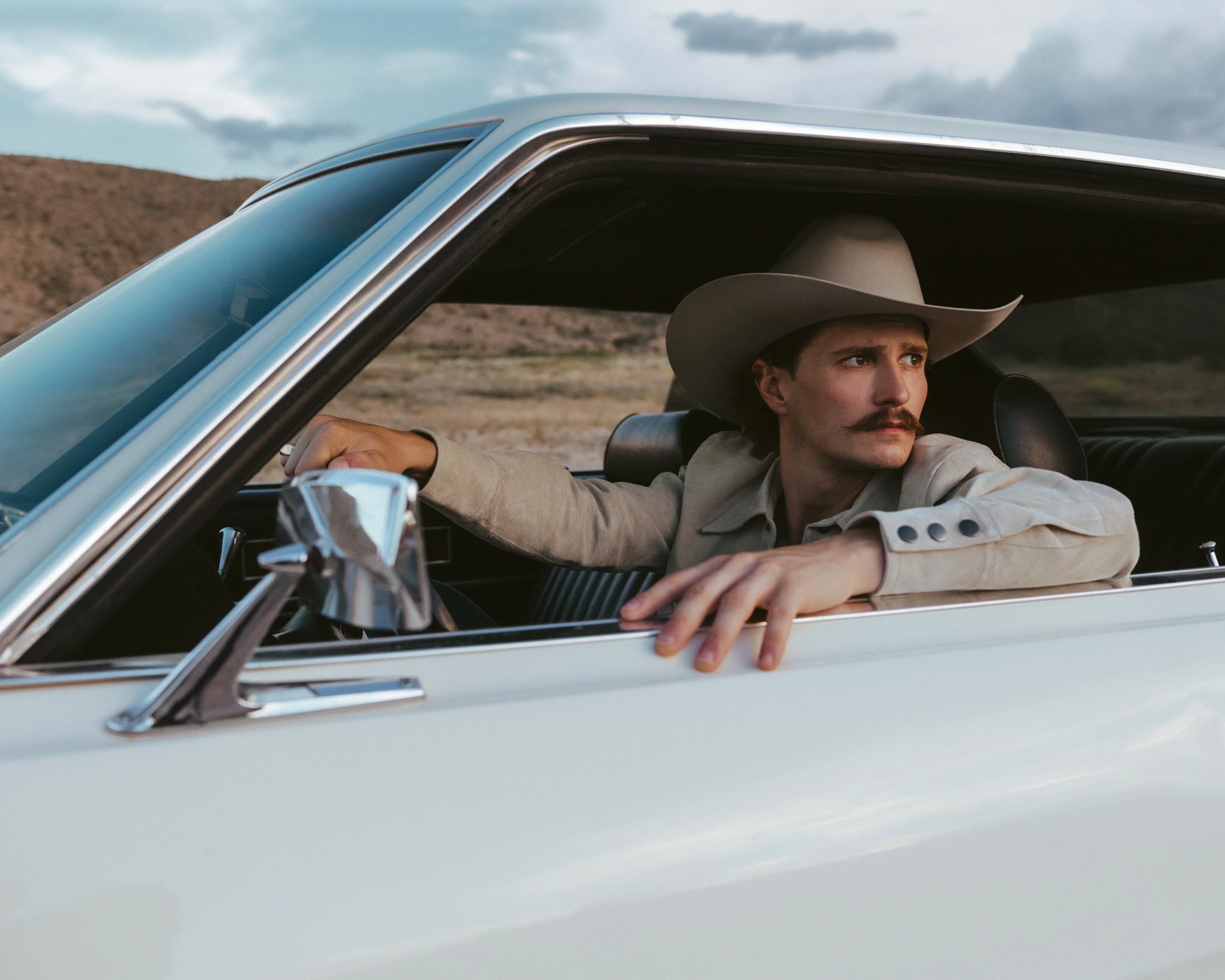 Braxton Keith at the wheel of a white vintage car in a cream felt hat and suede jacket, his arm resting on the open window.