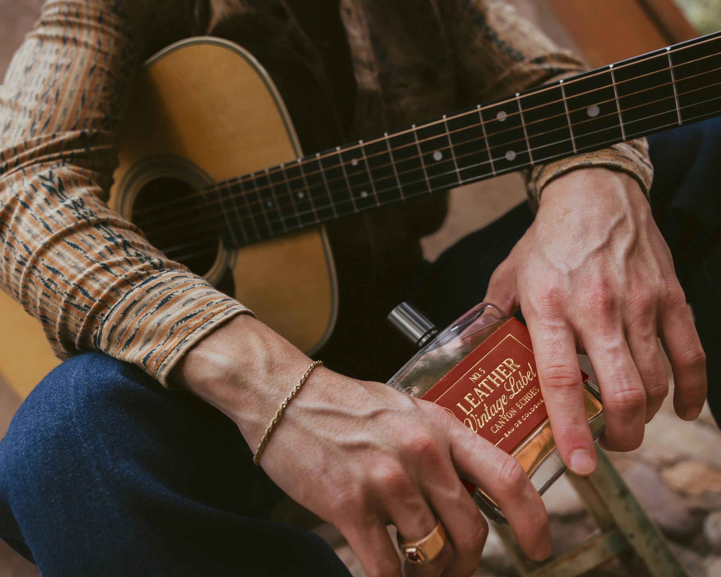Close on hands over an acoustic guitar, a gold bracelet and ring, turning the bottle with its red suede label in the light.
