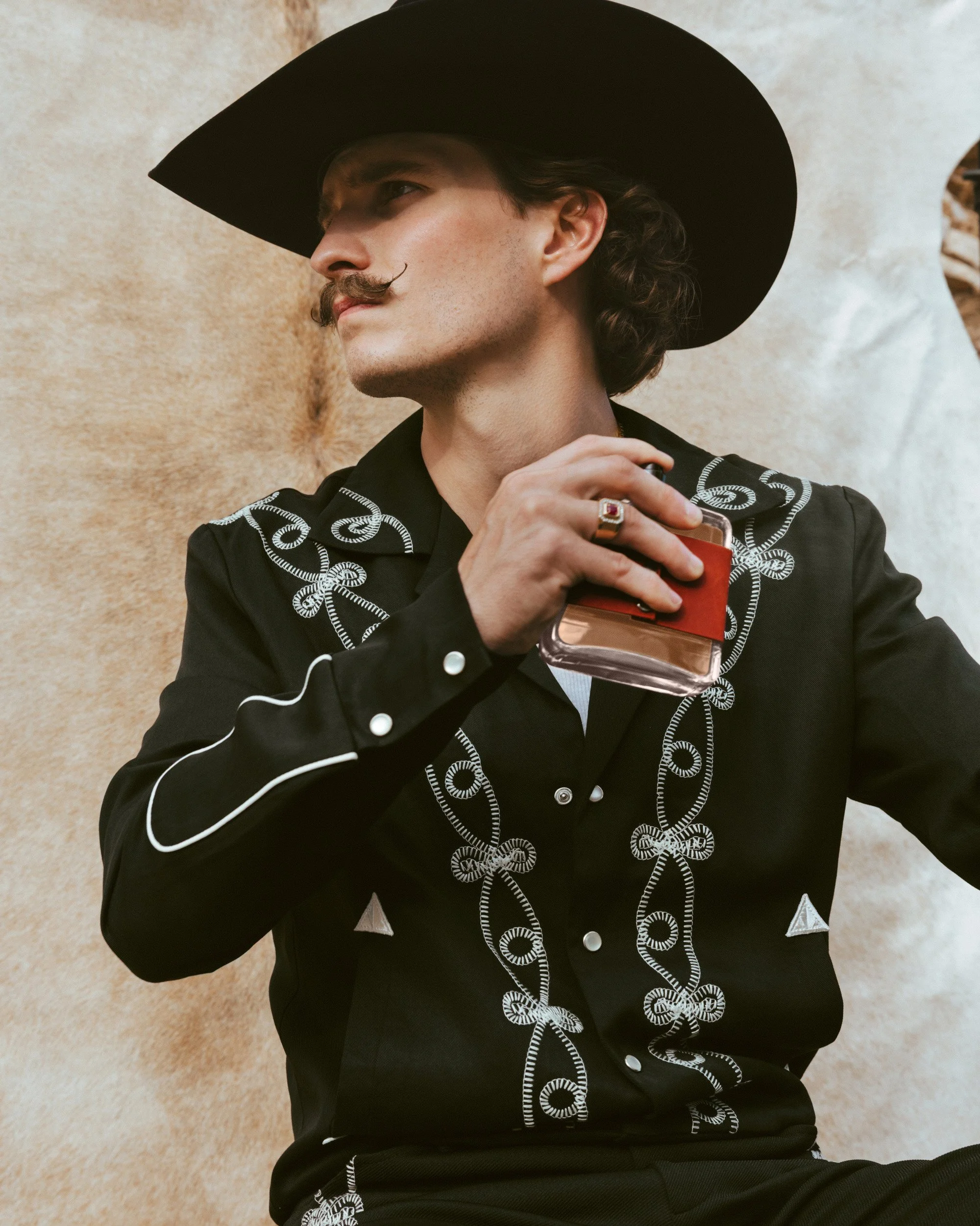 Braxton Keith in a black hat and black embroidered shirt, lifting the bottle in a ringed hand against a cowhide backdrop.