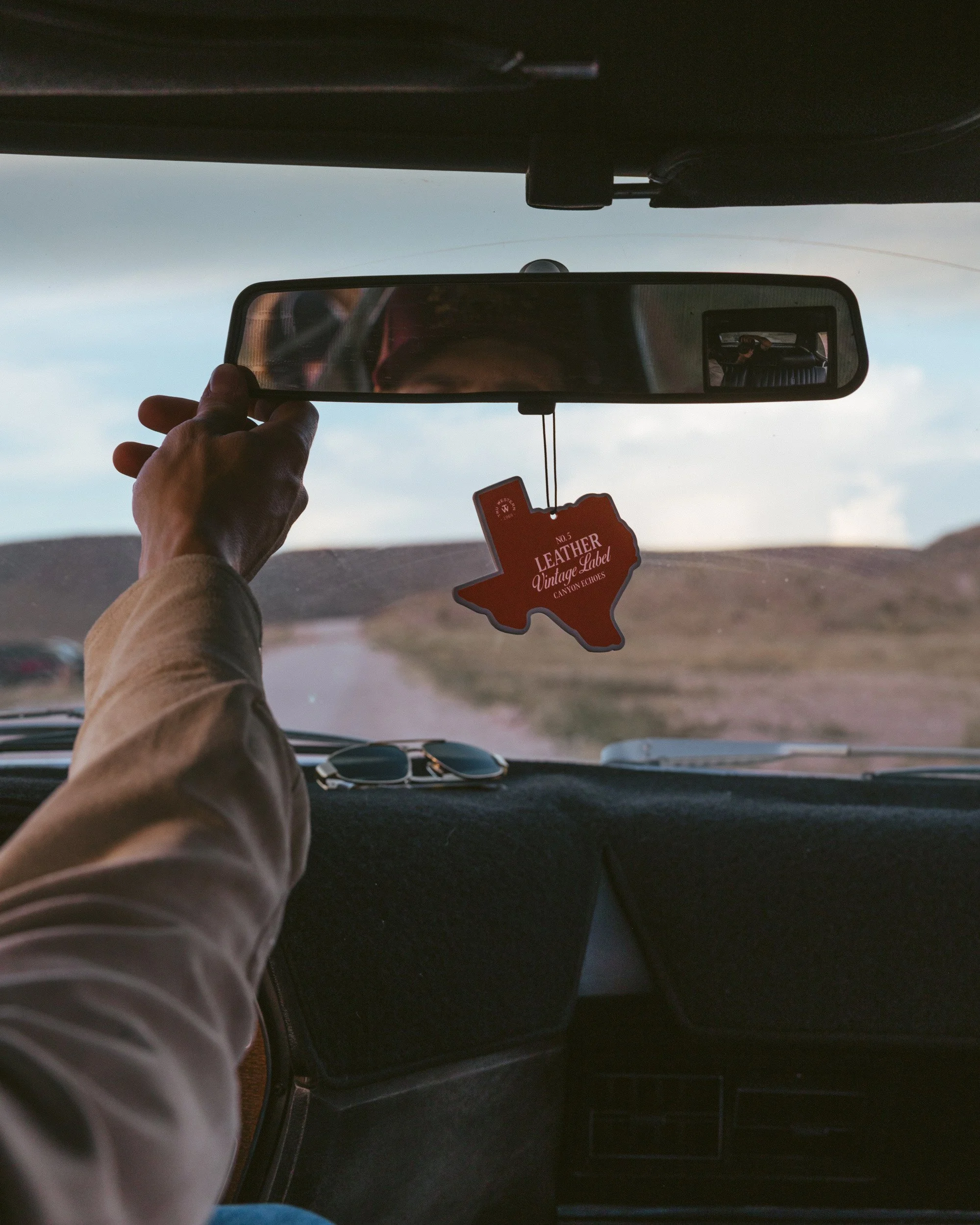 A hand reaches for the rearview mirror, a Texas-shaped Leather Vintage Label freshener swinging over the desert road.