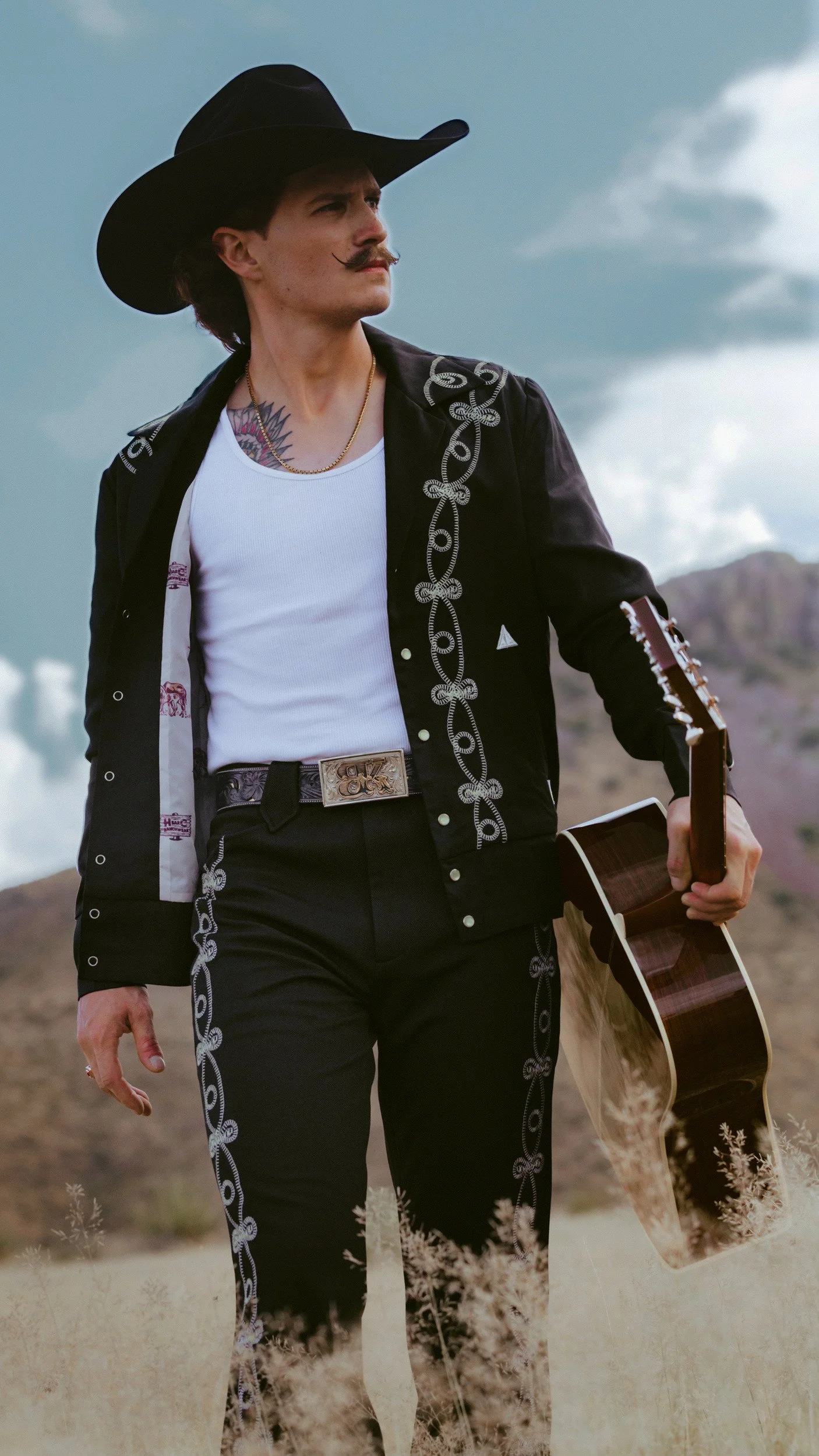 Braxton Keith walking through tall grass with his guitar, black hat, silver embroidered suit open over a white tank top.