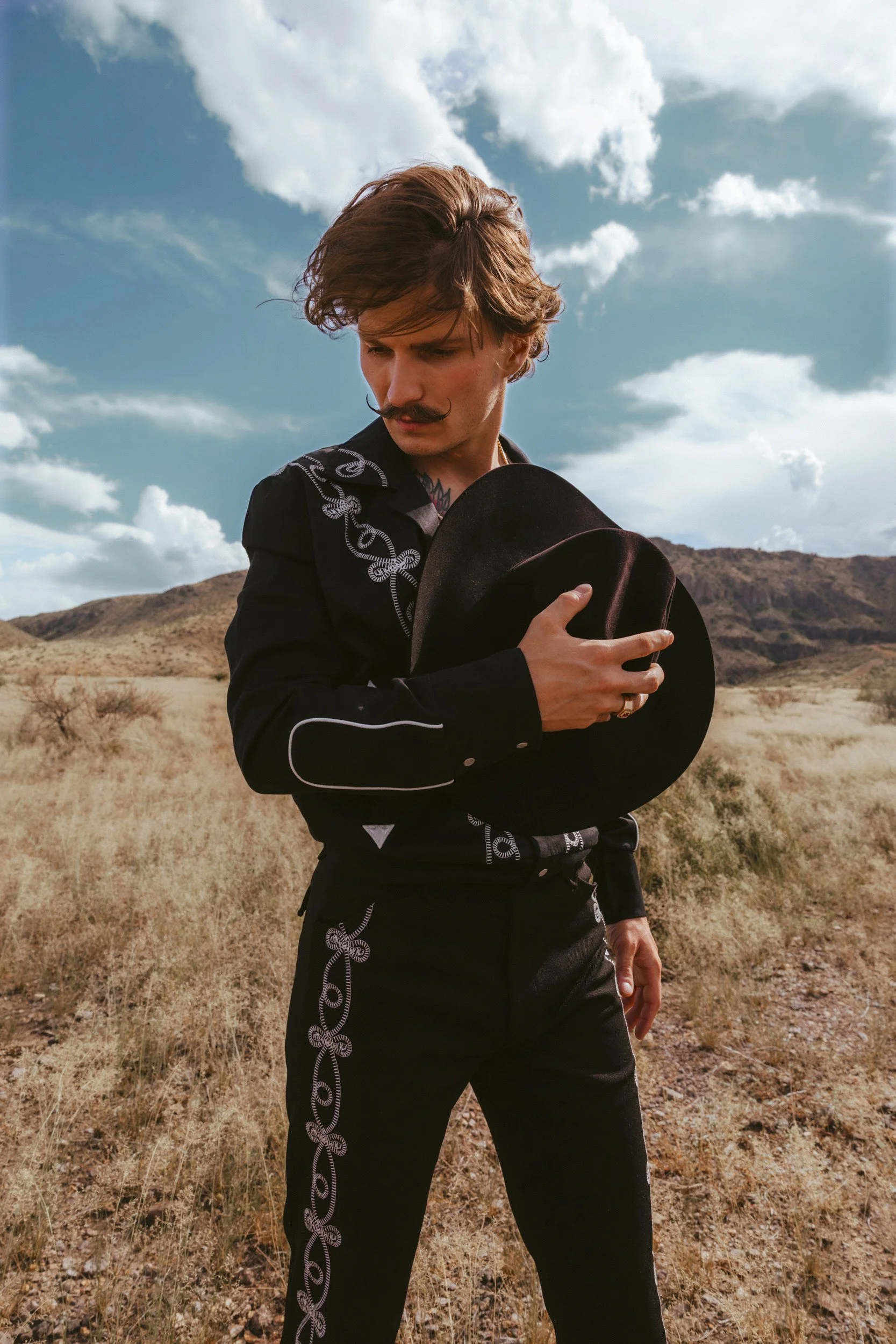 Braxton Keith in a black suit embroidered in silver, head bowed, holding his black hat to his chest in the open desert.