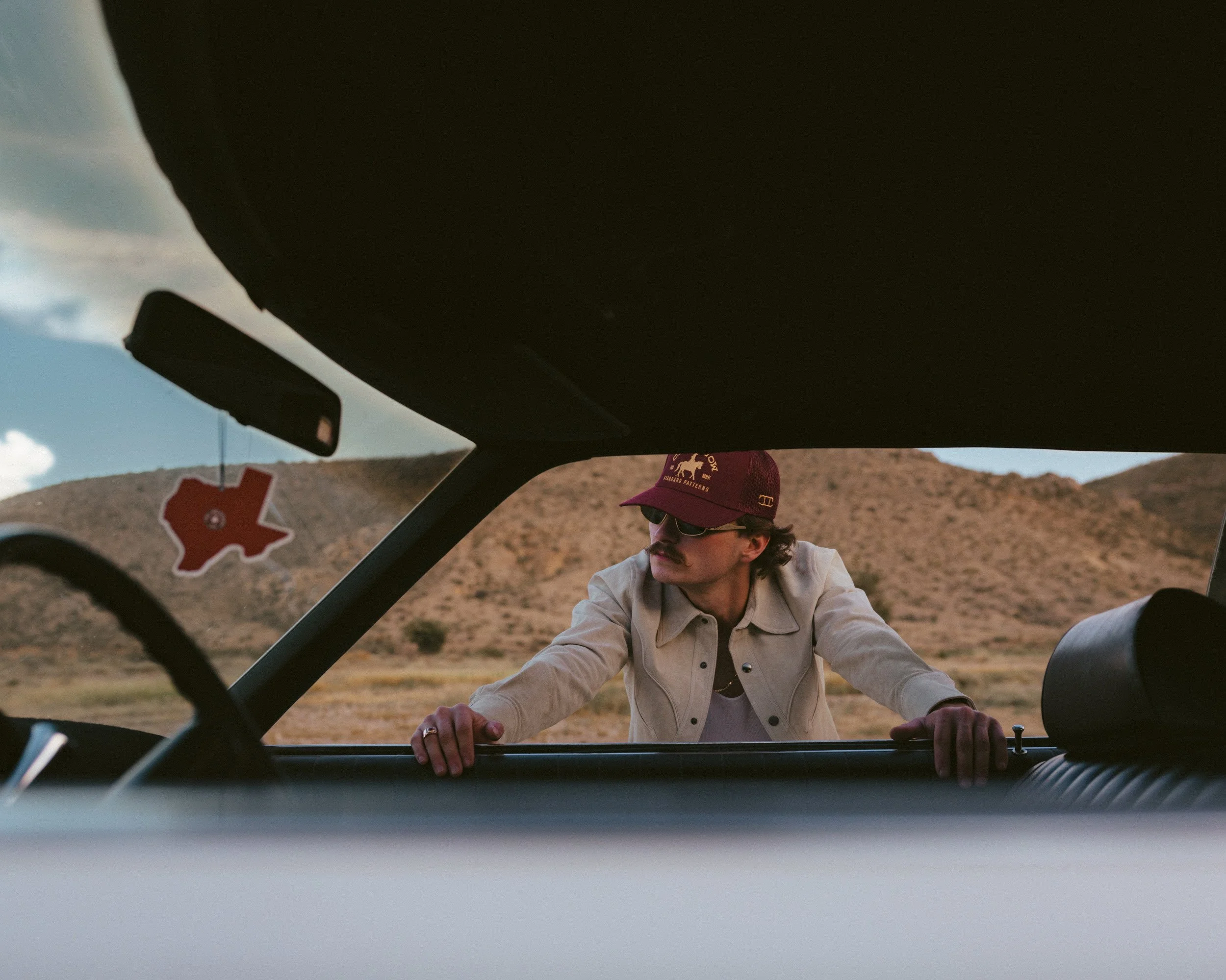 Through the windshield of a white car, Braxton Keith in a red cap leans on the hood beneath a hanging Texas freshener.
