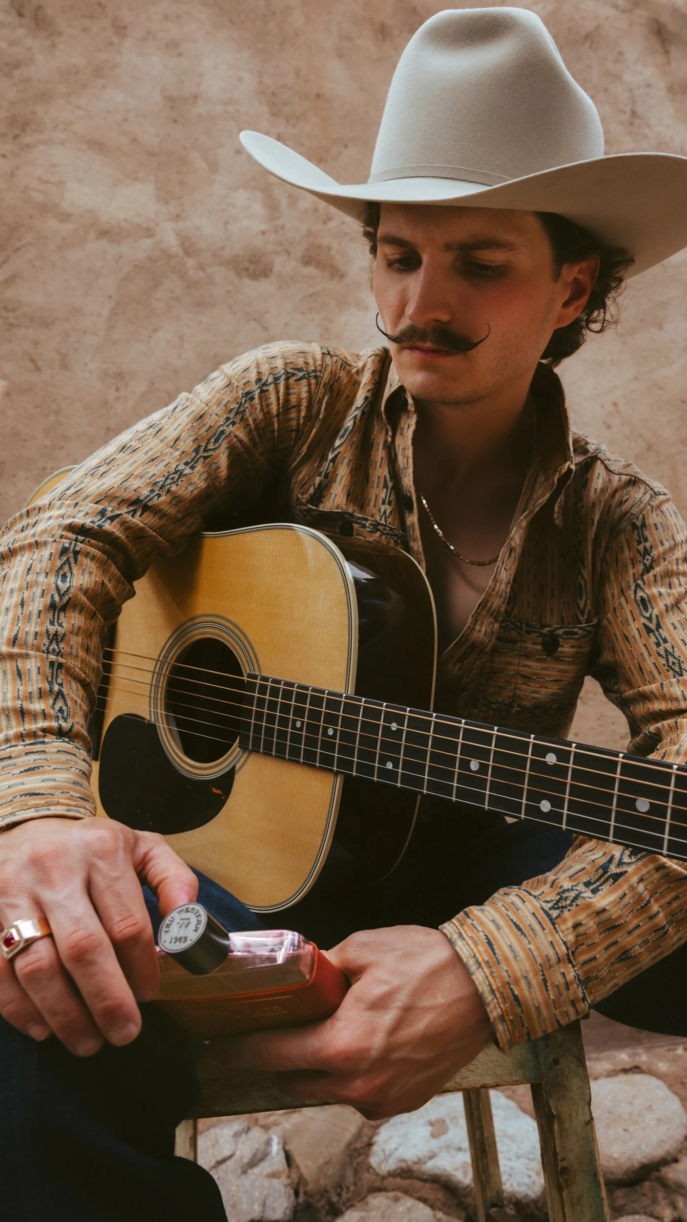 Braxton Keith in a white felt hat and patterned western shirt, seated with his guitar and the bottle in his ringed hands.