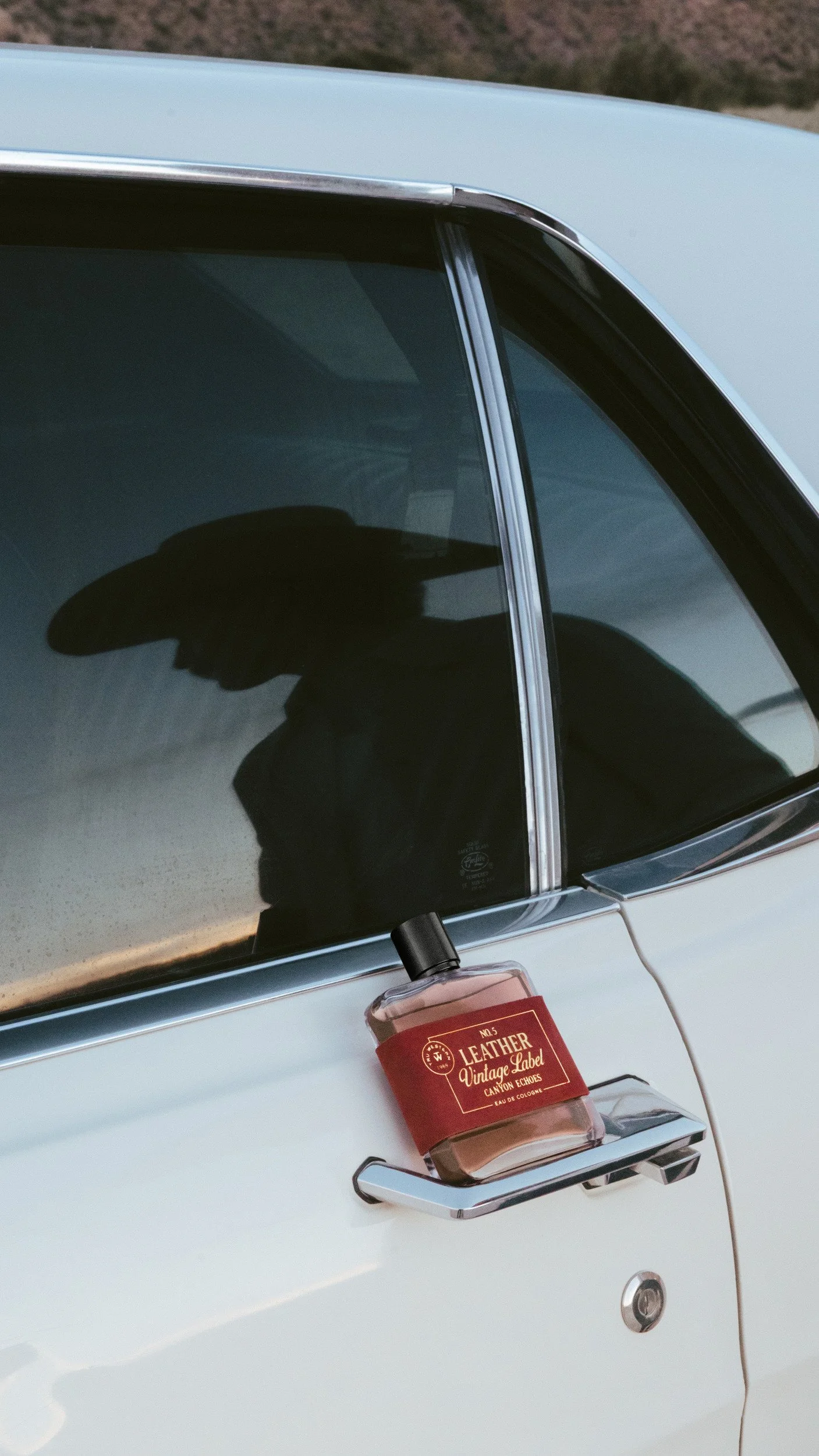 The bottle hooked on the chrome door handle of a white car, a cowboy hat silhouetted in the window glass above it.