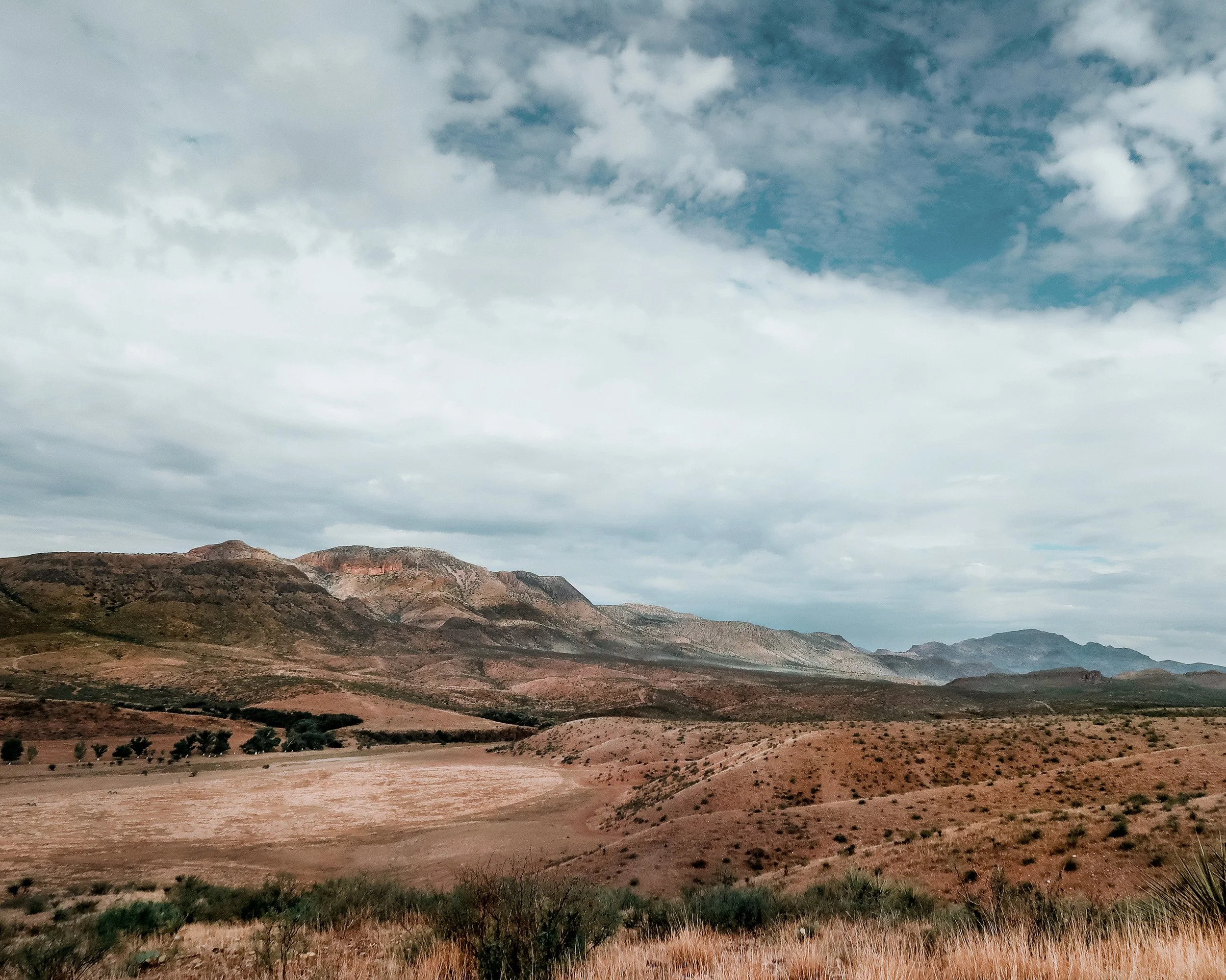 A wide West Texas valley under a sky of moving cloud, red and ochre hills folding away toward the mountains on the horizon.