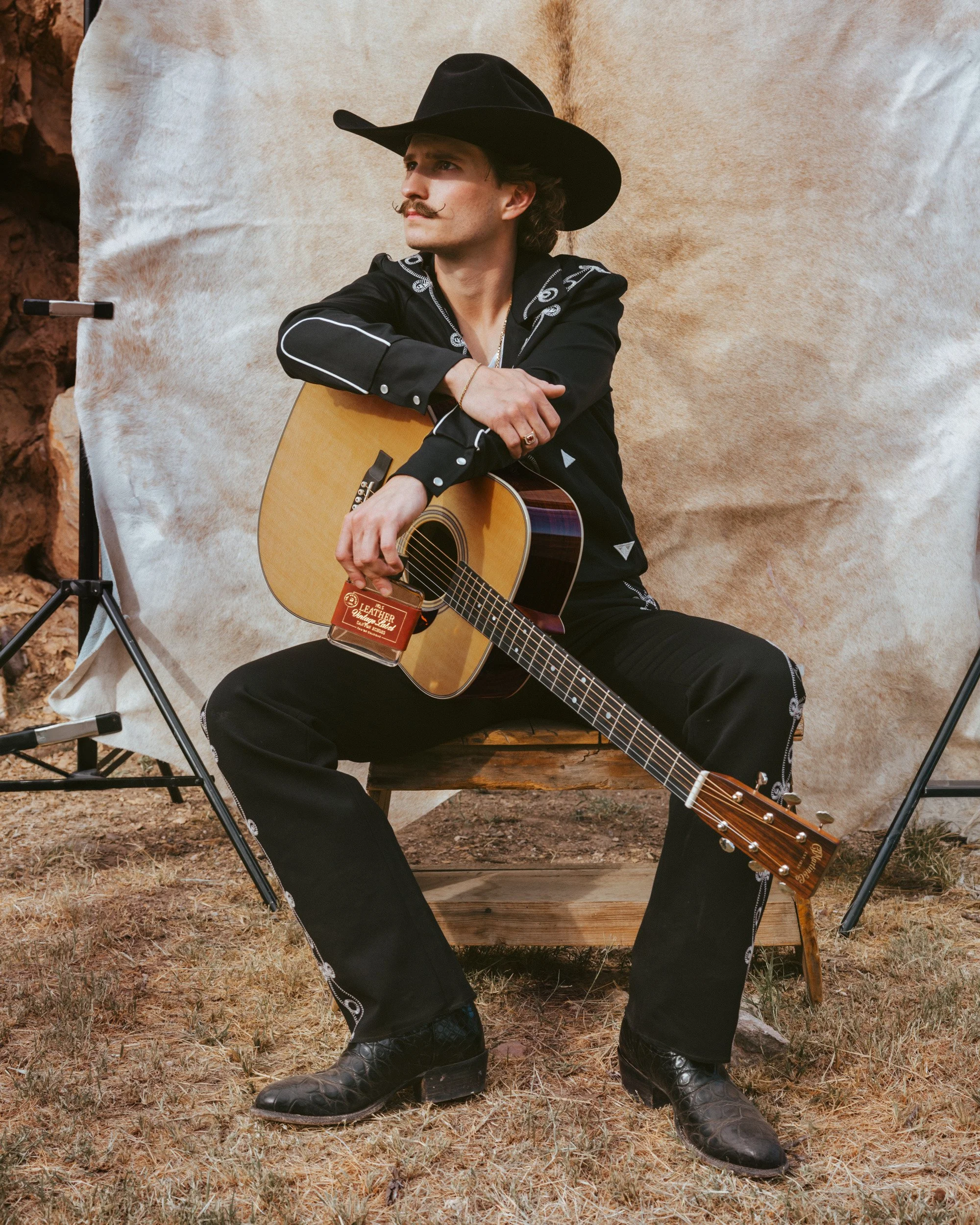 Braxton Keith on a wooden crate at a cowhide backdrop on set, guitar and bottle in hand, black hat and black boots.