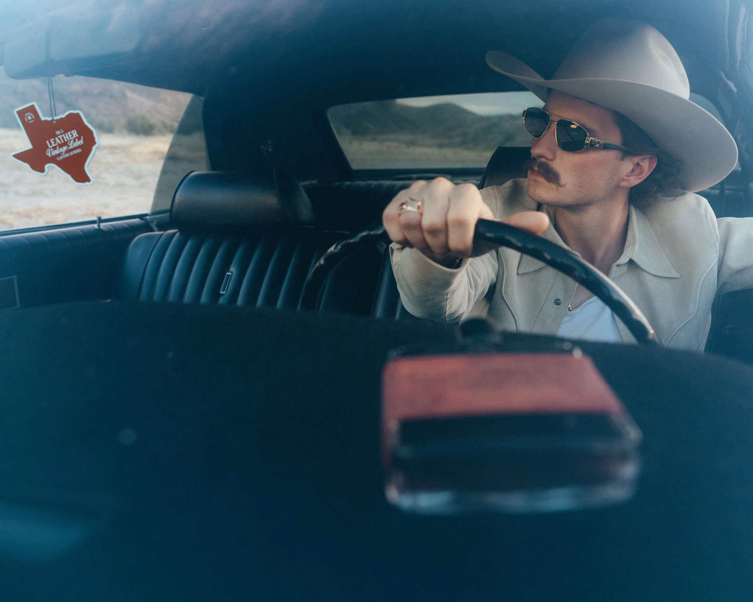 Braxton Keith driving in aviator sunglasses and a cream hat, a Texas freshener in the window and the bottle on the dash.