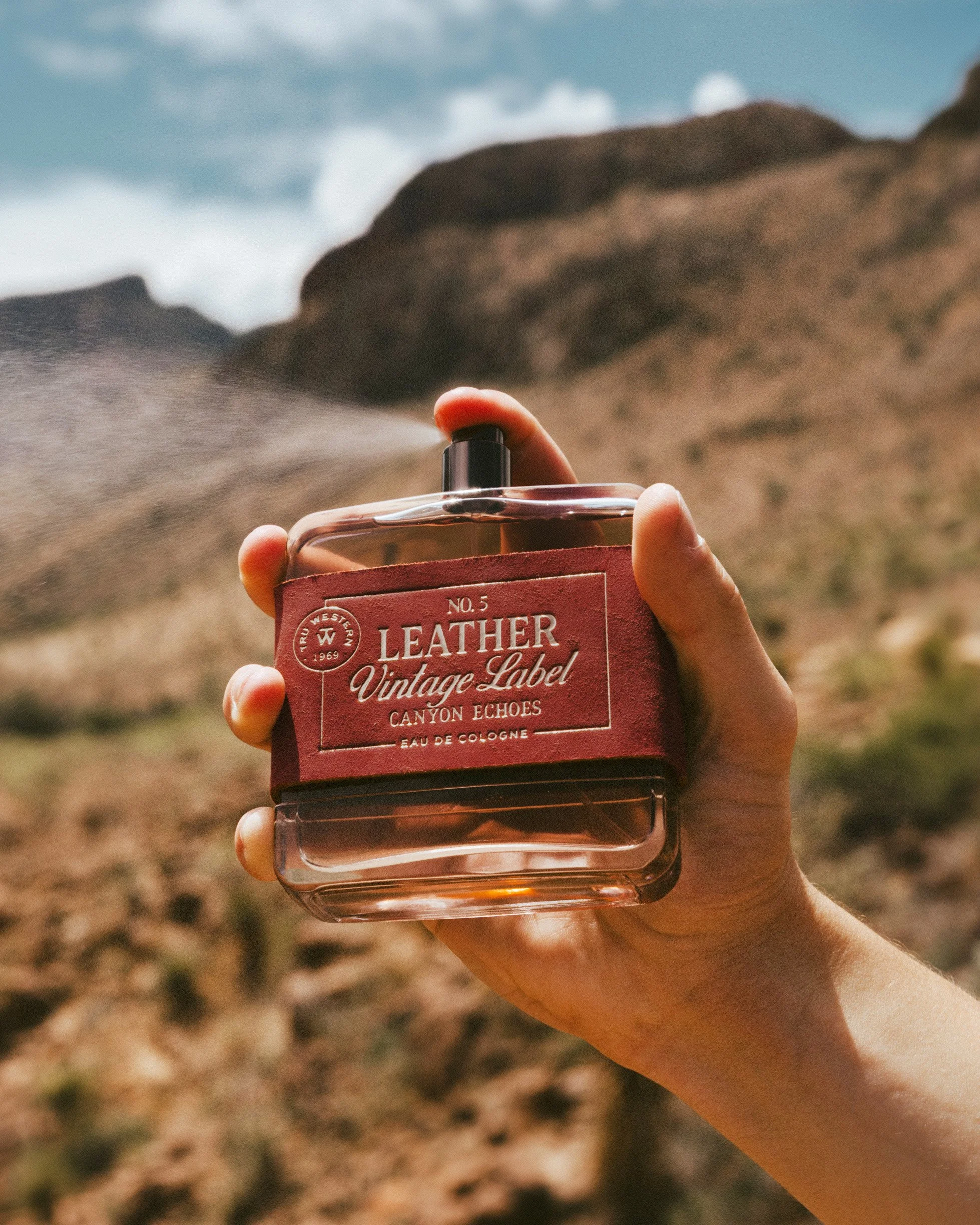 A hand raises No. 5 Leather Vintage Label: Canyon Echoes against the desert hills, a fine mist of spray drifting off it.