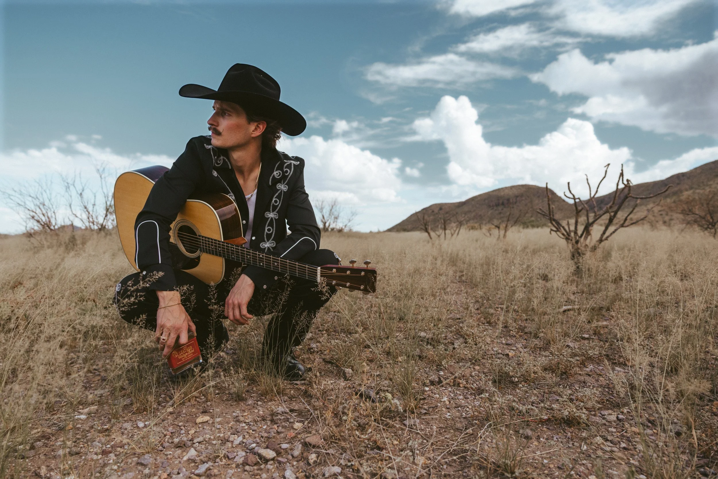 Braxton Keith crouched in dry desert grass in a black embroidered suit and black hat, guitar on his knee, the bottle in hand.