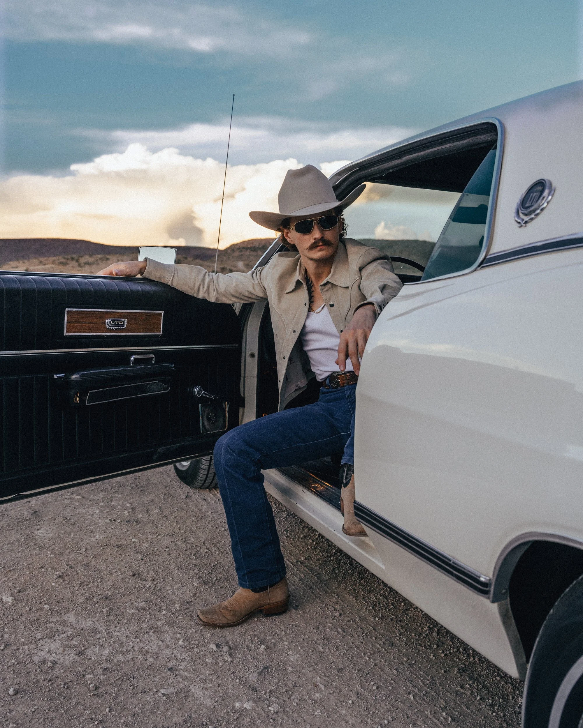 Braxton Keith stepping out of a white car in a cream hat, sunglasses and jeans, one boot on the gravel, clouds lit behind.