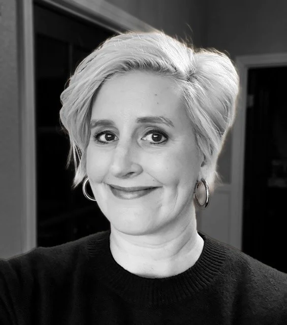 Black and white photo of a woman with short, wavy hair, smiling, wearing hoop earrings and a dark sweater.
