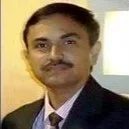 A young woman with shoulder-length black hair smiling brightly, wearing a blue top, surrounded by green plants in a bright indoor setting.