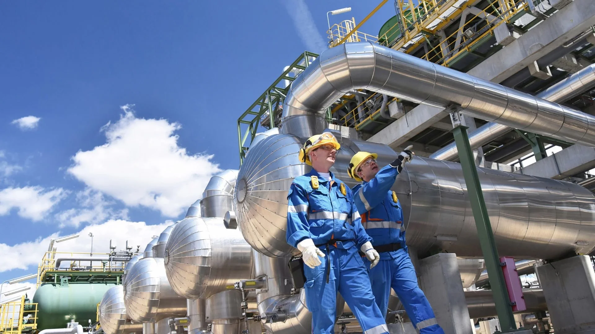 Two workers in blue uniforms and yellow protective helmets inspecting industrial pipes at an oil or gas facility under a bright blue sky.