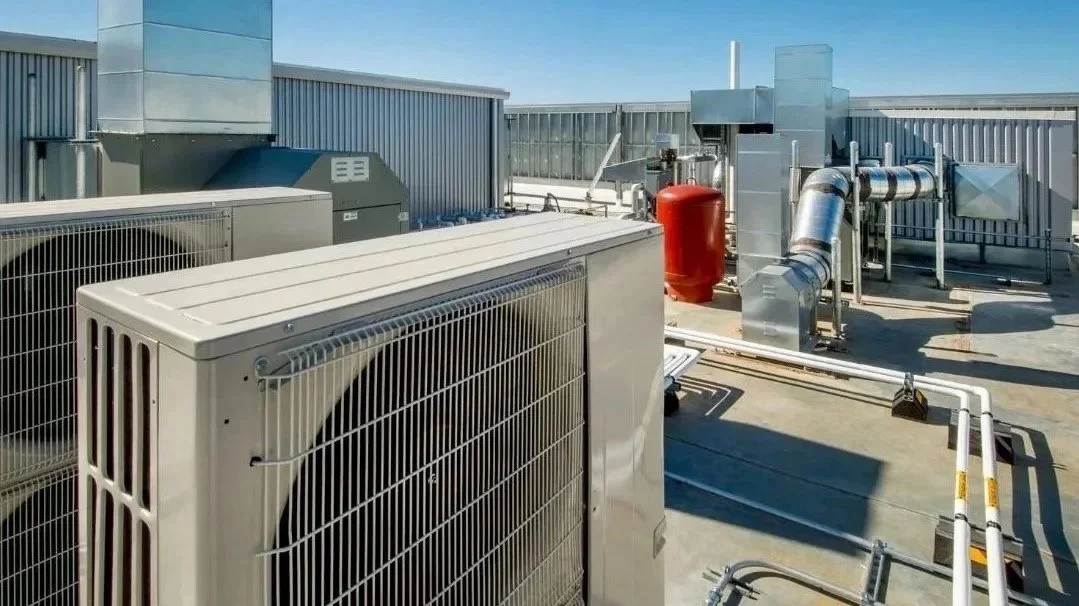 Rooftop HVAC equipment including air conditioning units, ductwork, pipes, and a red fire suppression tank on a building's rooftop under a clear blue sky.