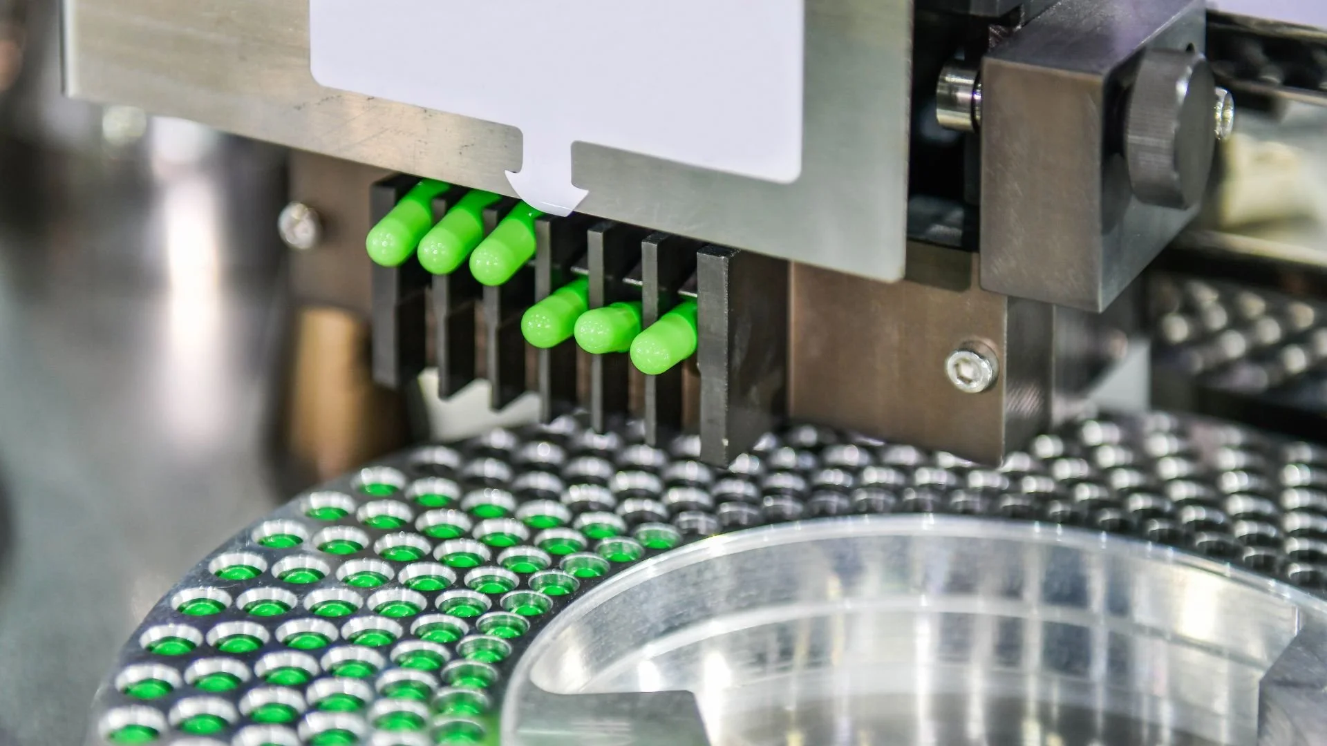 Close-up of an automated machine placing green capsules into a circular tray with multiple holes.