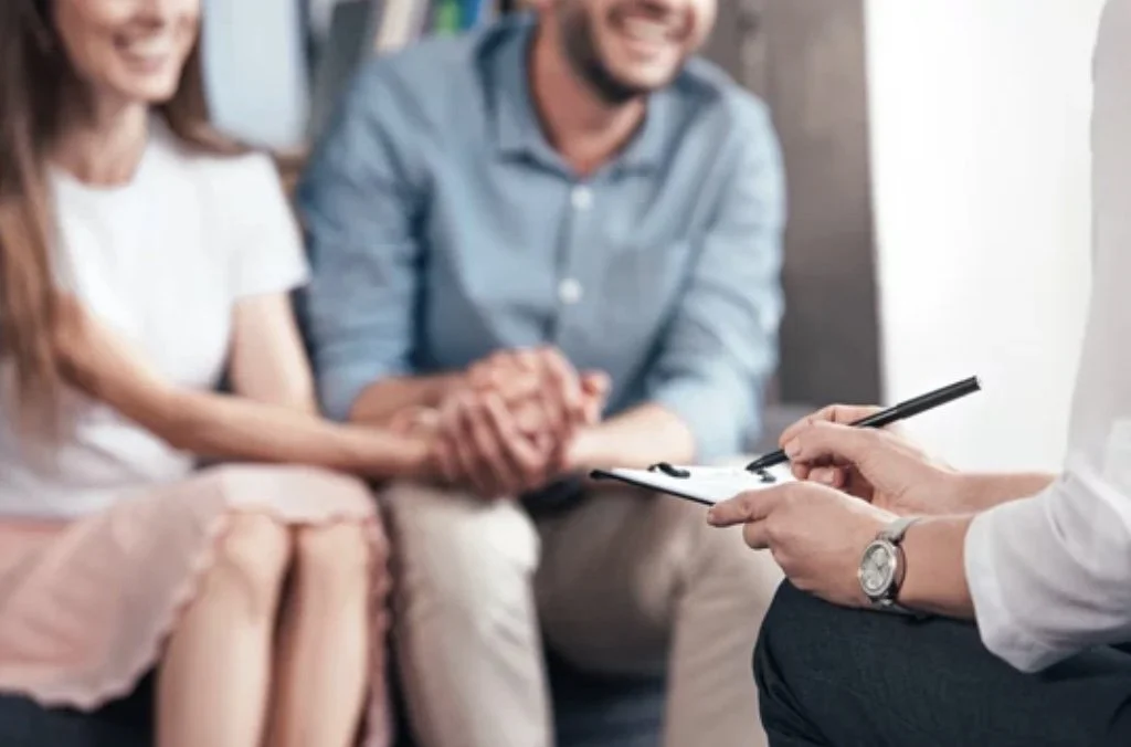 A couple sitting on a couch during a therapy session, holding hands, with a therapist taking notes on a clipboard.