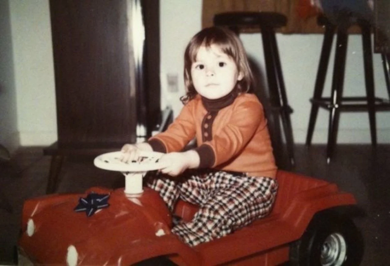 A young girl with brown hair wearing an orange jacket and checkered pants is sitting in a red toy car with a black gift bow on the front. She is indoors near a wooden table and chairs.