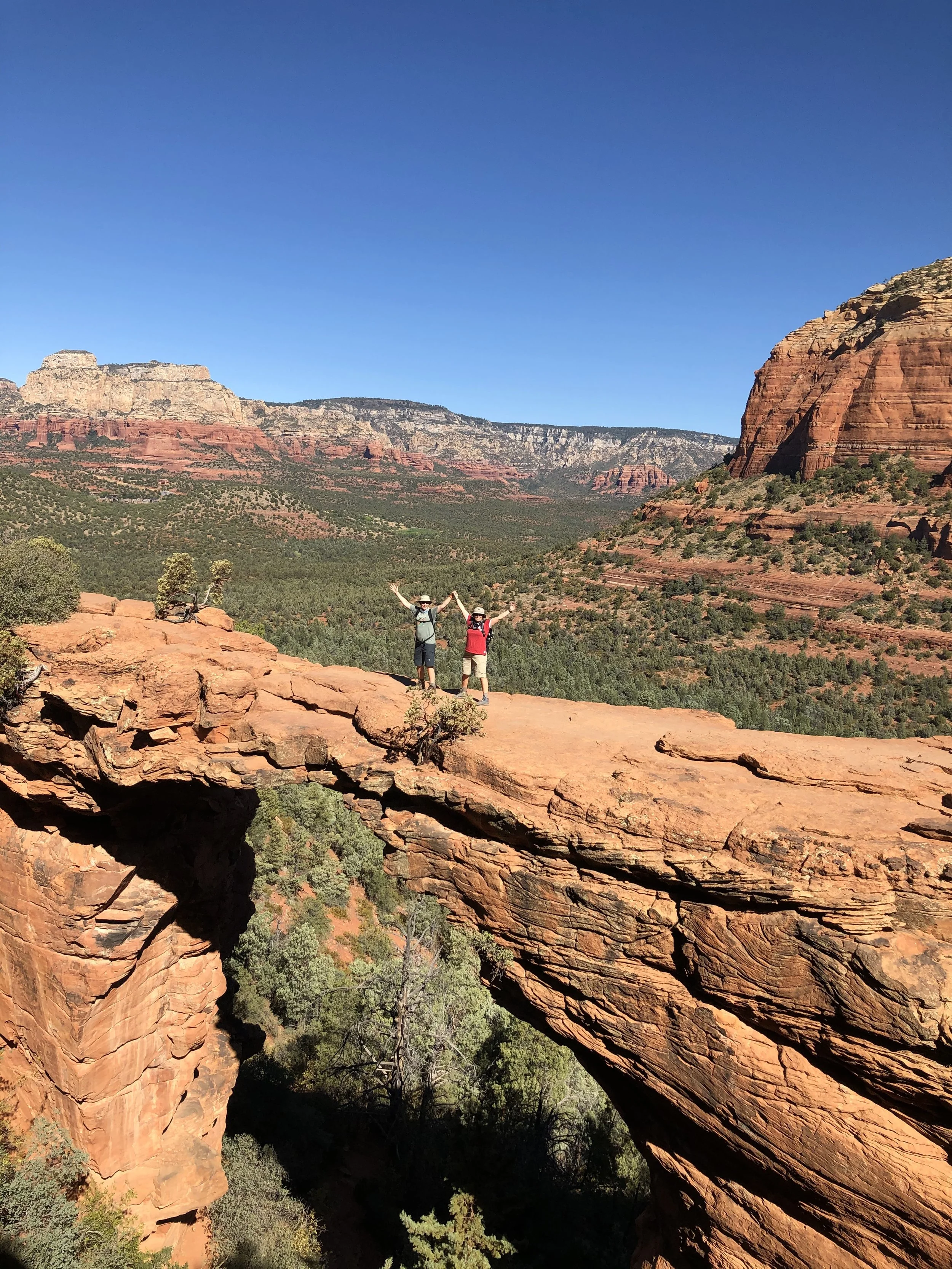 Two hikers standing on a natural rock arch in a desert landscape with red rock formations and green vegetation under a clear blue sky.