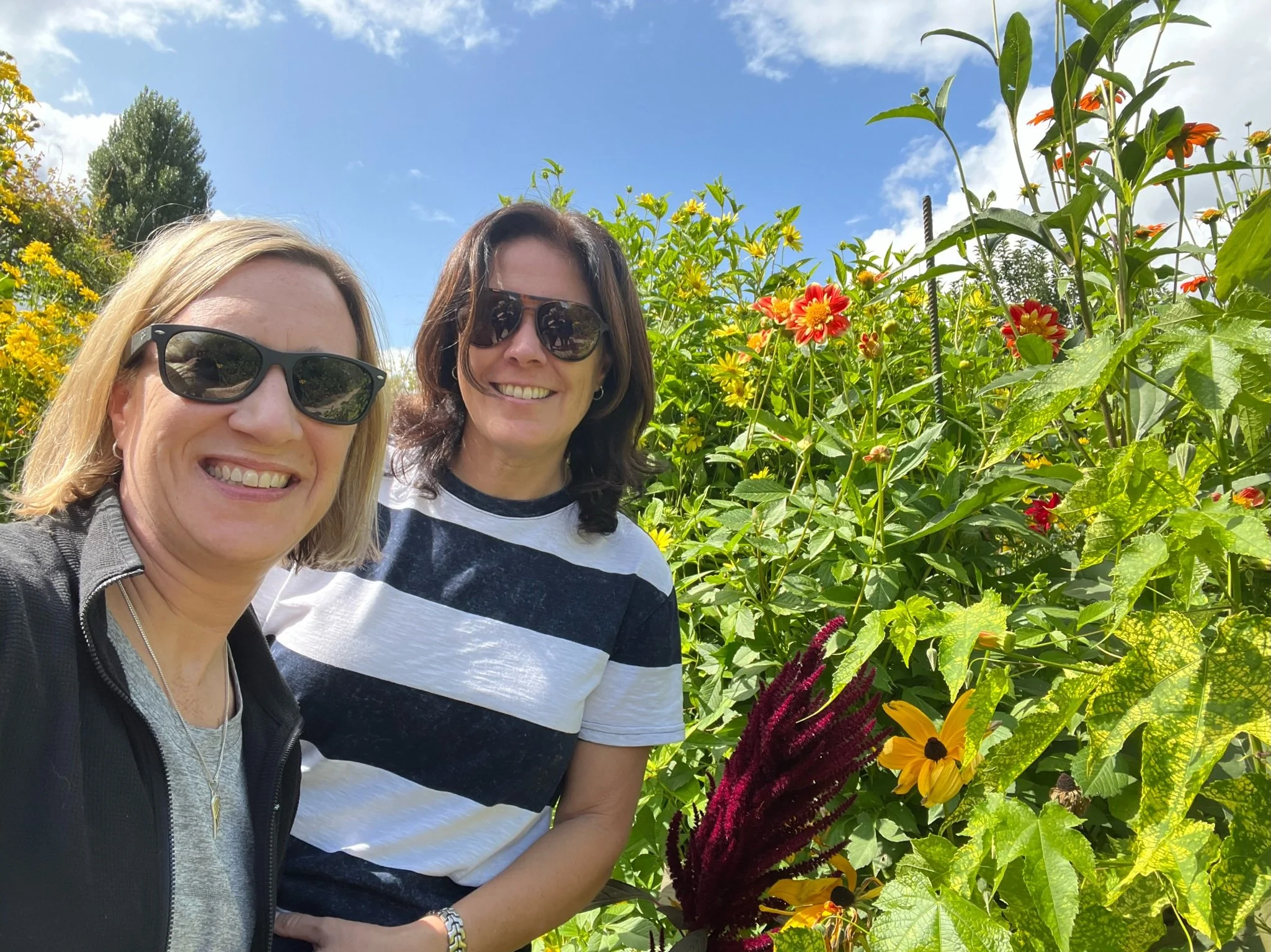 Two women wearing sunglasses smiling in a garden with colorful flowers and green foliage under a partly cloudy blue sky.