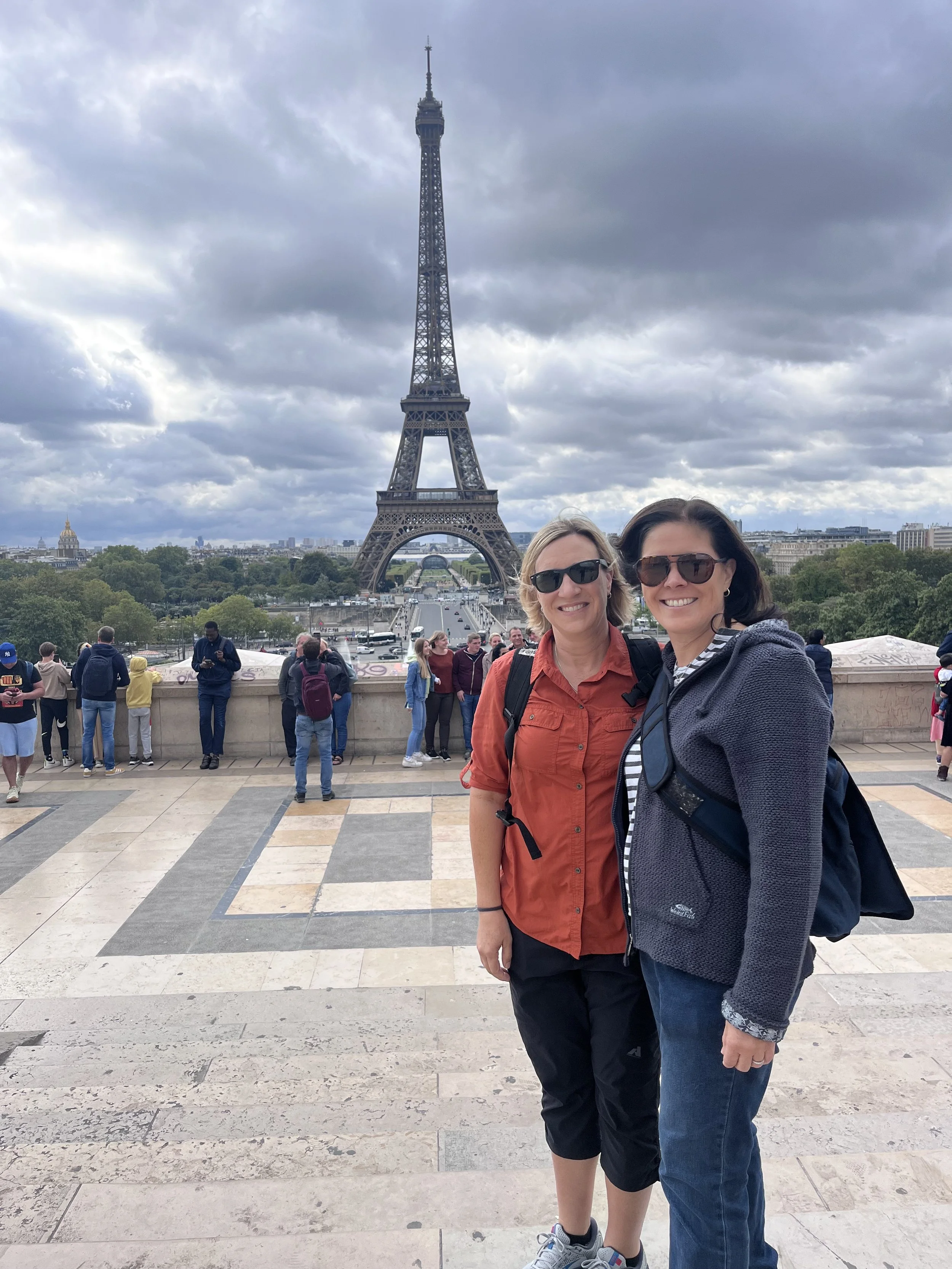 Two women smiling and posing in front of the Eiffel Tower in Paris, with cloudy skies and tourists in the background.