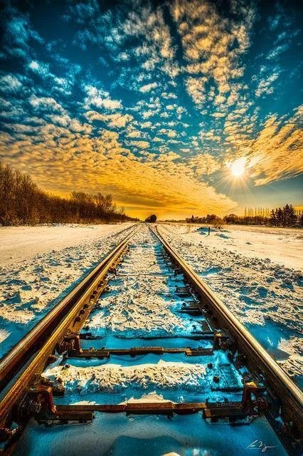 Train tracks in a snowy landscape with a vibrant sunset sky and scattered clouds.