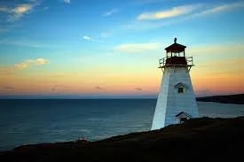 A white lighthouse on a grassy hill overlooking the ocean at sunset.