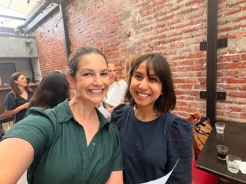 Two women smiling and taking a selfie in a brick-walled room with other people in the background.