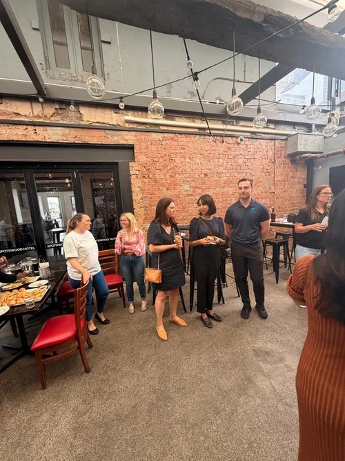 Group of five people standing and talking in a restaurant or cafe with exposed brick wall and string lights hanging from the ceiling.