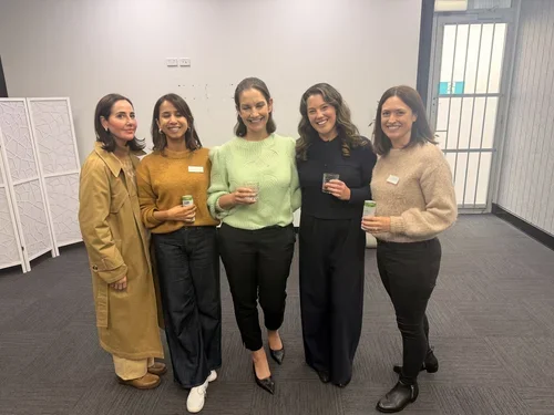 Five women standing together in an indoor setting, holding drinks, dressed in semi-formal attire, smiling for the photo.