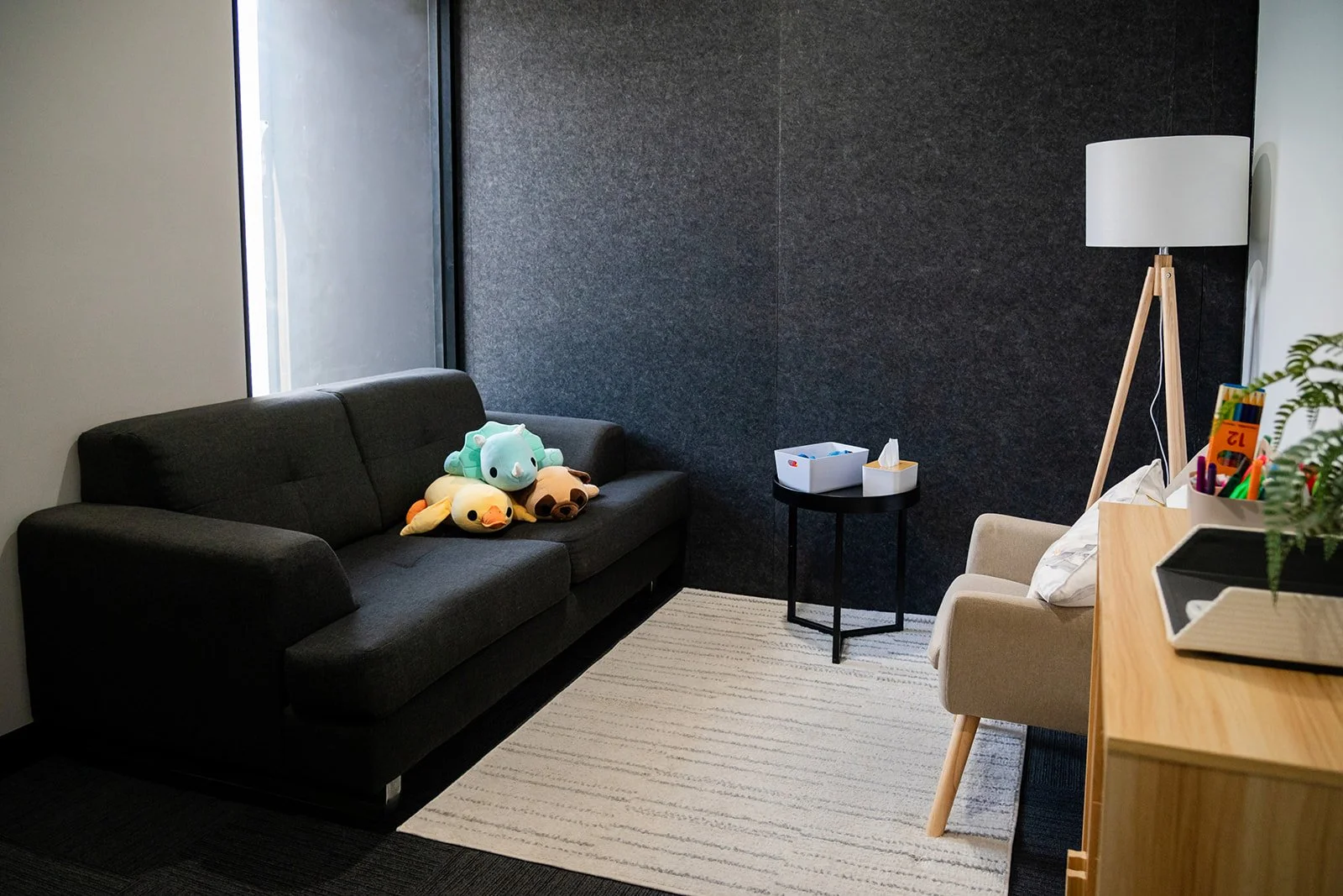 Living room with a black sofa holding plush toys, a small beige armchair with pillows, a black side table with tissues, a wooden desk with colored pens, a potted plant, and a tall white floor lamp. The room has a textured dark wall and a window letti
