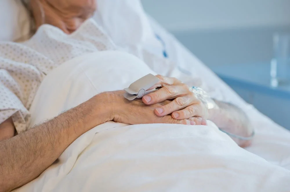 Close-up of a patient's hands resting on their stomach in a hospital bed, with one hand holding an oxygen monitor.
