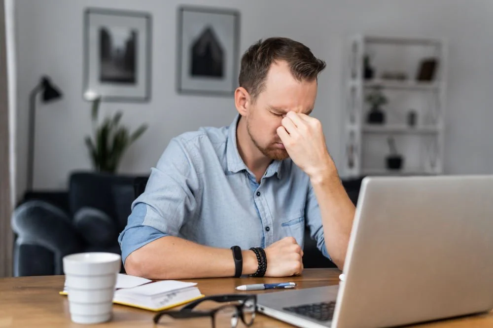 Man sitting at a desk with a laptop, pinching his nose in frustration, surrounded by notebooks and a coffee mug.