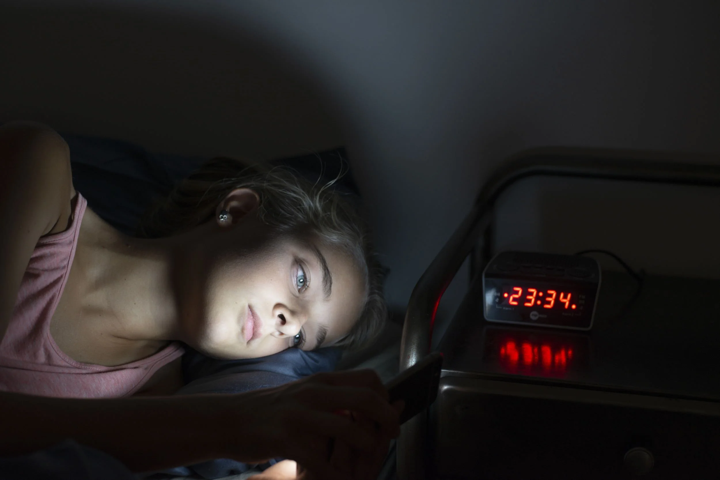 A young girl lying down next to an alarm clock displaying the time 11:34. The girl has light brown hair, blue eyes, and is wearing a pink sleeveless top. She appears to be resting on a dark surface in a dimly lit room.
