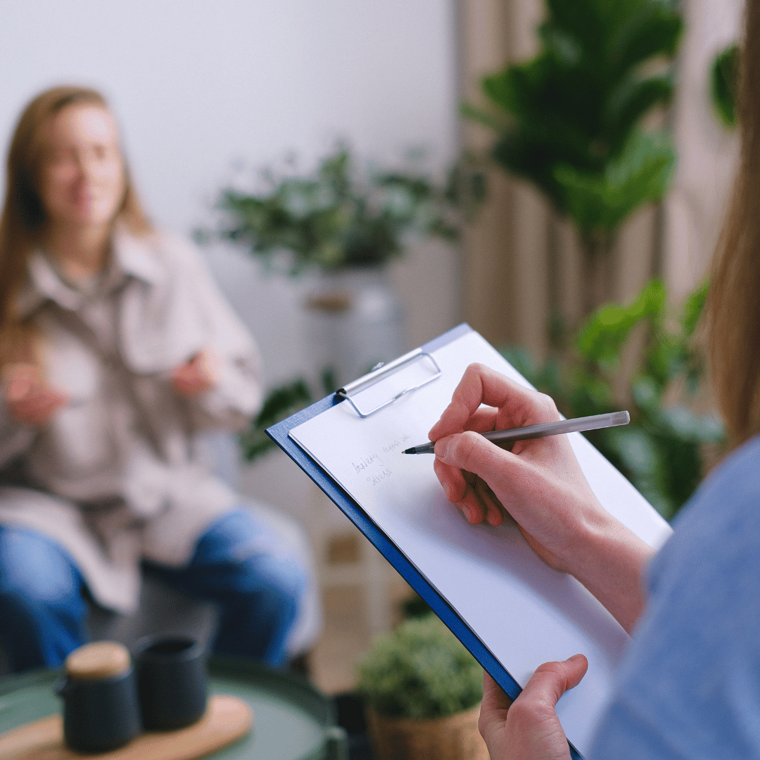 Therapist taking notes on a clipboard during a counseling session with a patient sitting in a cozy, plant-filled room.