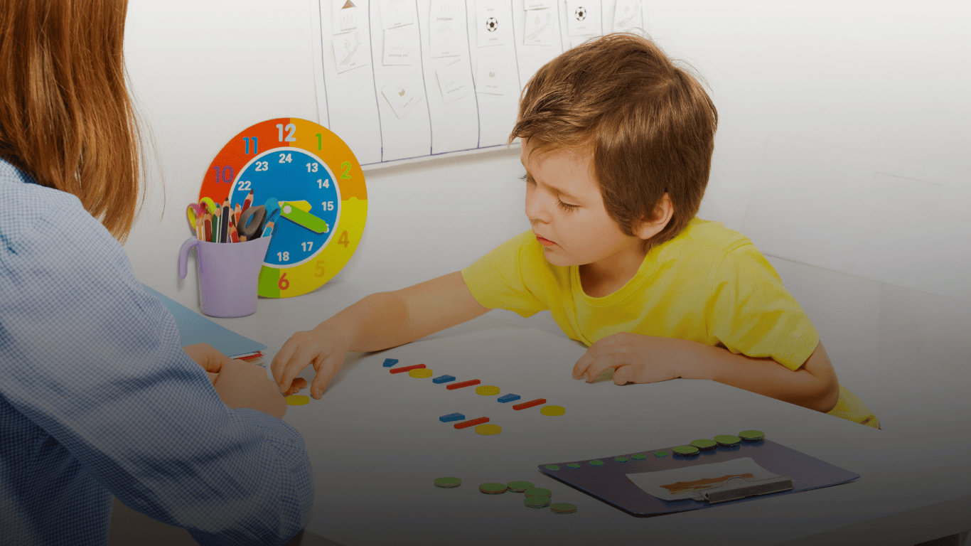 A young boy wearing a yellow shirt is playing a sorting game with colorful shapes on a white table, supervised by an adult.