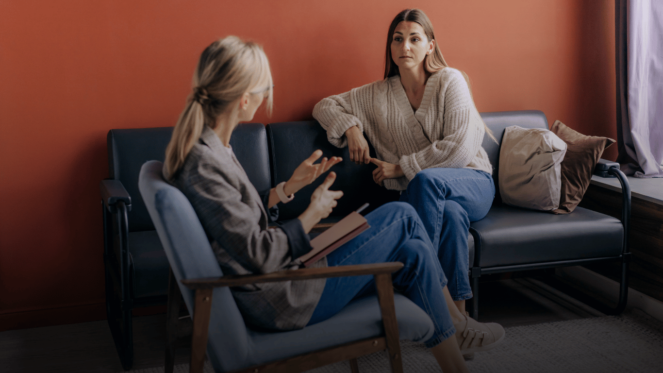 Two women having a conversation in a living room with orange walls, sitting on a black sofa and a blue armchair. One woman with blonde hair is gesturing while holding a notebook, and the other woman with brown hair is listening attentively.