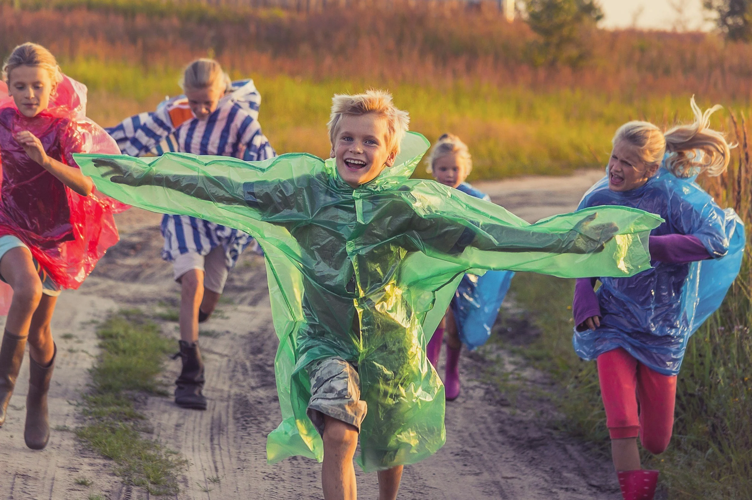 Children running outdoors in a field dressed in colorful raincoats during sunset.