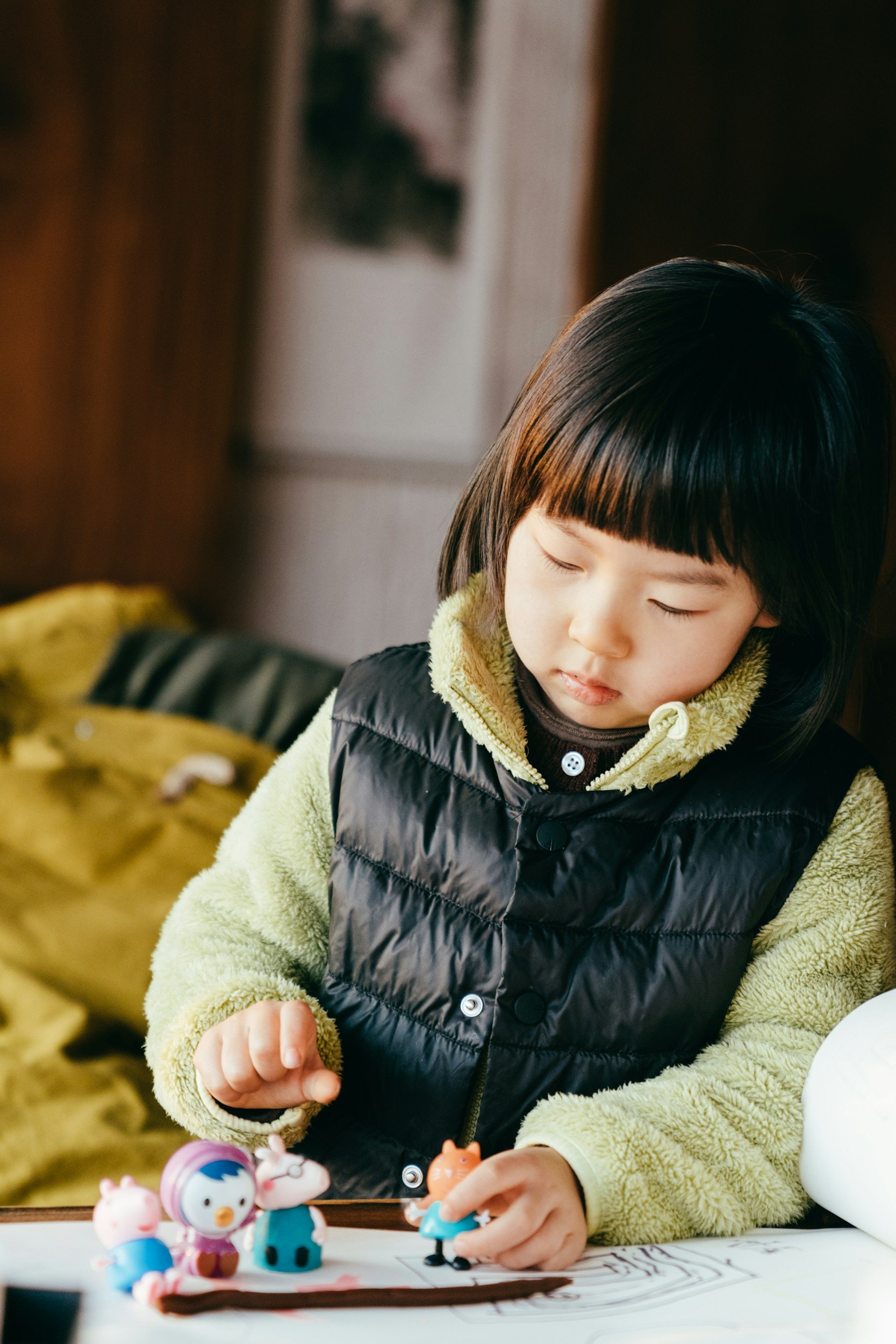 A young girl with dark hair and bangs, wearing a light green sweater and a black puffer vest, is playing with small colorful toy figures on a table. She is focused on the toys, and there are toy figurines and coloring pages on the table in front of her.