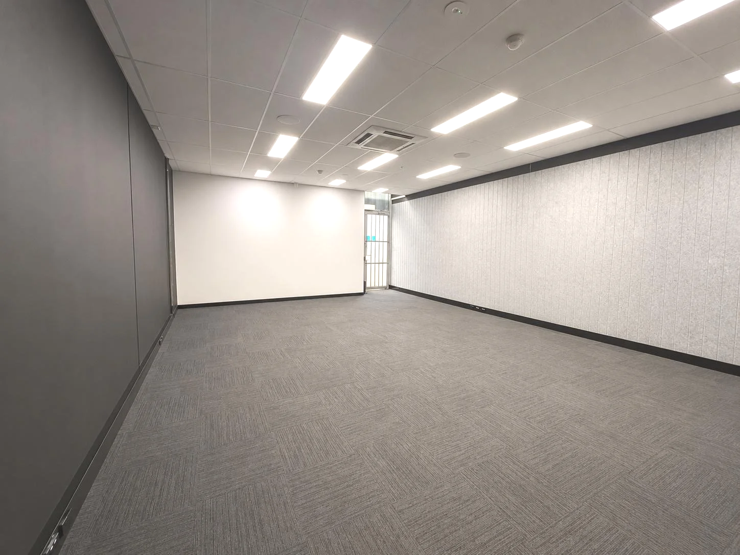 Empty office space with gray walls, gray carpet, white ceiling with bright fluorescent lights, and a door at the back.