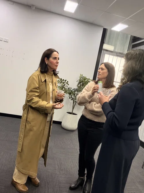 Three women standing and talking in an office, each holding drinks.