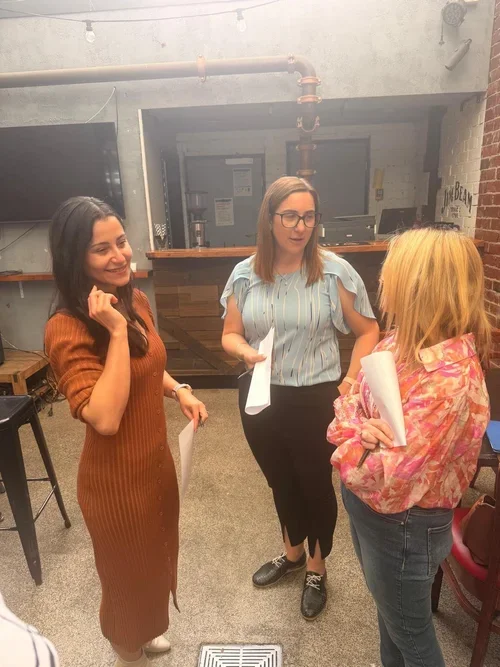 Three women are having a conversation in a modern, industrial-style room with exposed pipes, a brick wall, and a wooden counter. One woman is wearing a brown dress, the second is wearing a striped blouse, and the third is in a pink patterned jacket. They are holding papers and seem engaged in a discussion.