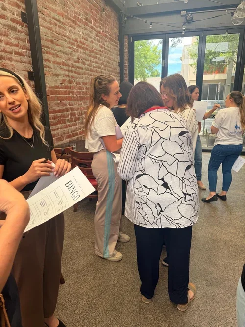Group of people standing in line inside a room with large glass windows, brick wall, and string lights, waiting to play bingo.