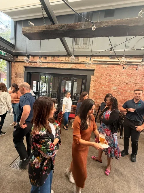 People socializing at an indoor gathering with exposed brick walls, string lights, and a large window door.