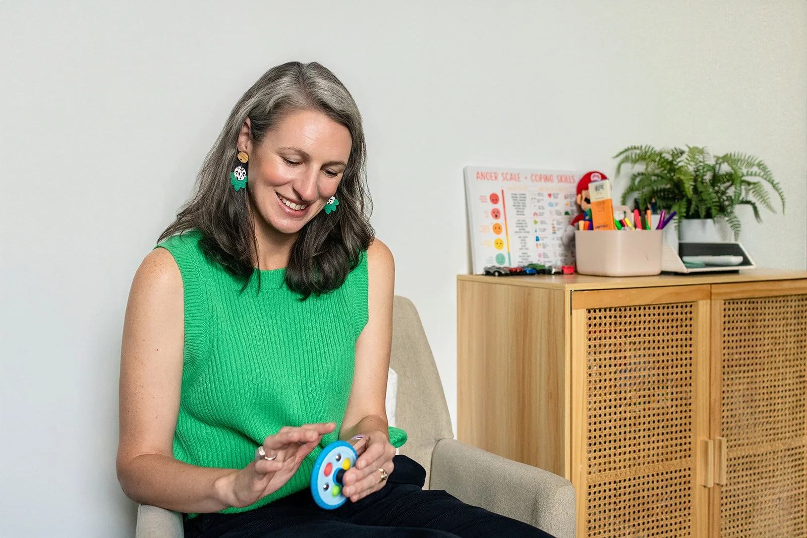 Woman in a green sleeveless top sitting on a beige armchair, smiling, holding a small handheld device with colorful buttons.