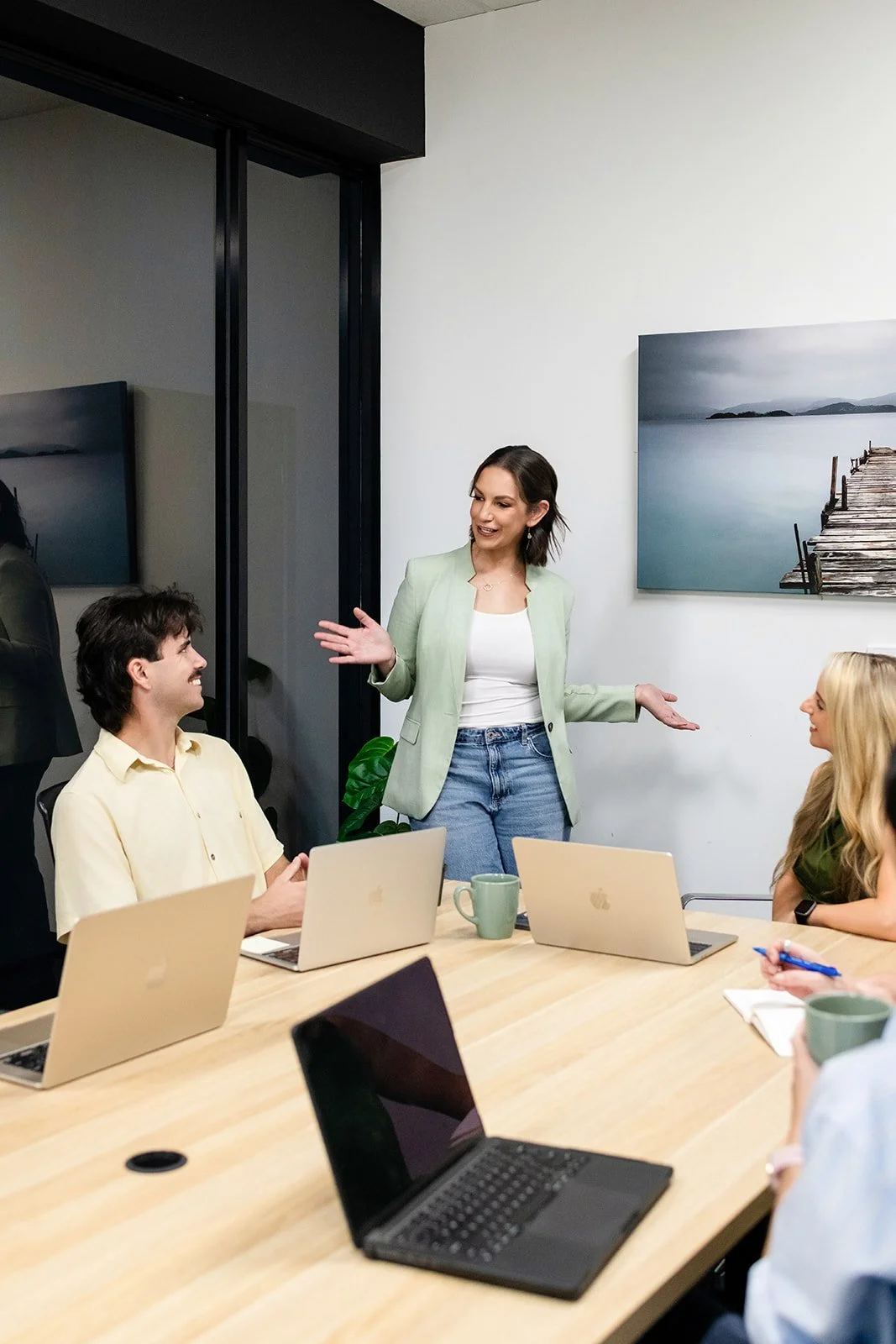 A woman standing and speaking to a group of people in a meeting room with laptops on the table and a landscape painting on the wall.