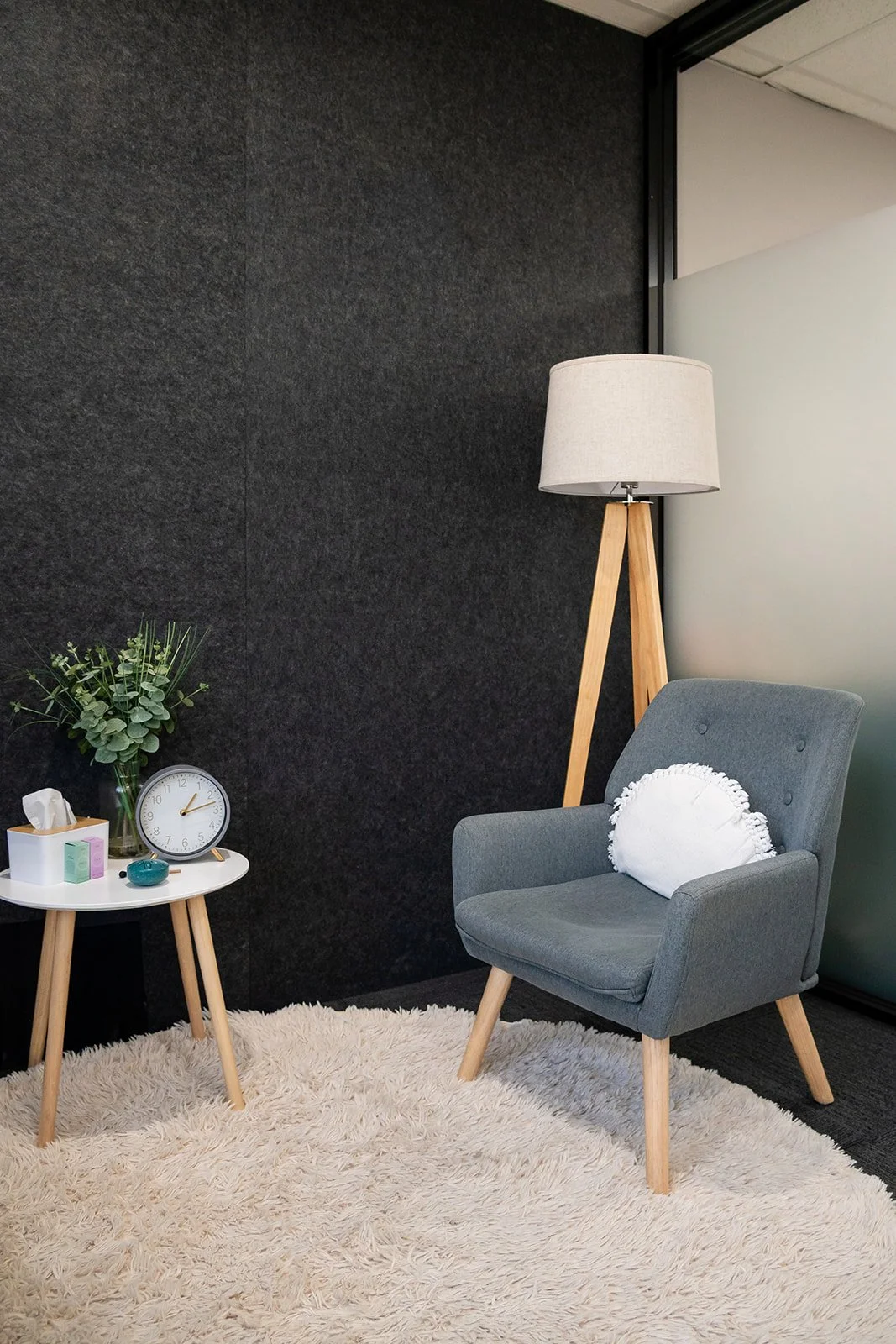 A cozy corner with a gray armchair and white fluffy rug, a small round side table with a clock, tissue box, decorative items, and a green plant, next to a tall wooden floor lamp with a beige lampshade against a black textured wall and frosted glass w
