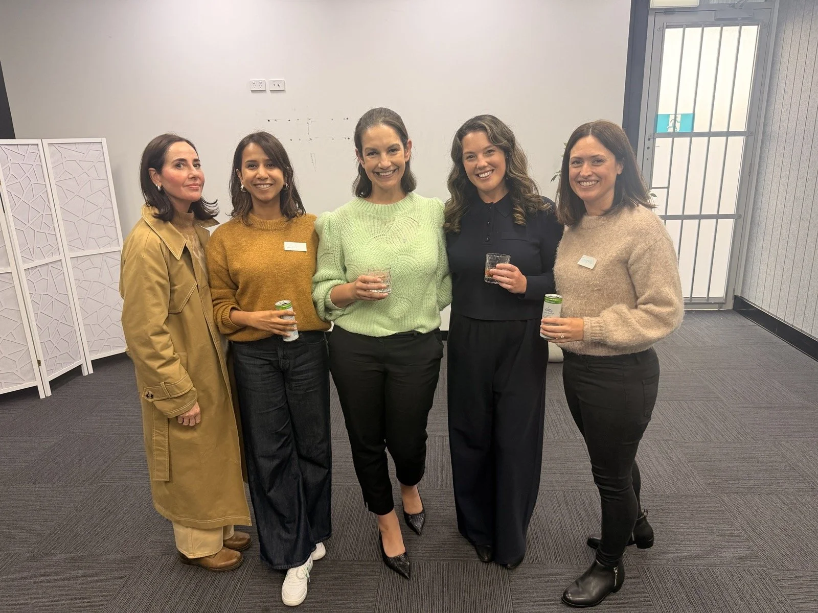 Group of five women standing together indoors, smiling, holding drinks and cans, dressed in casual and business casual clothing.